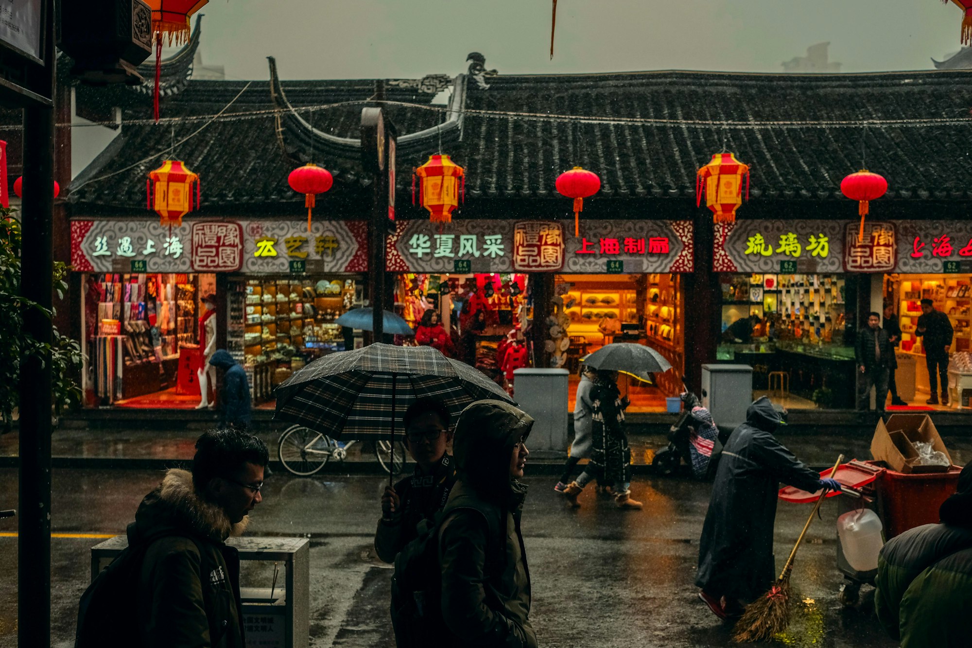 Shoppers with umbrellas pass lantern-hung Chinese shopfronts on a rainy street at dusk.
