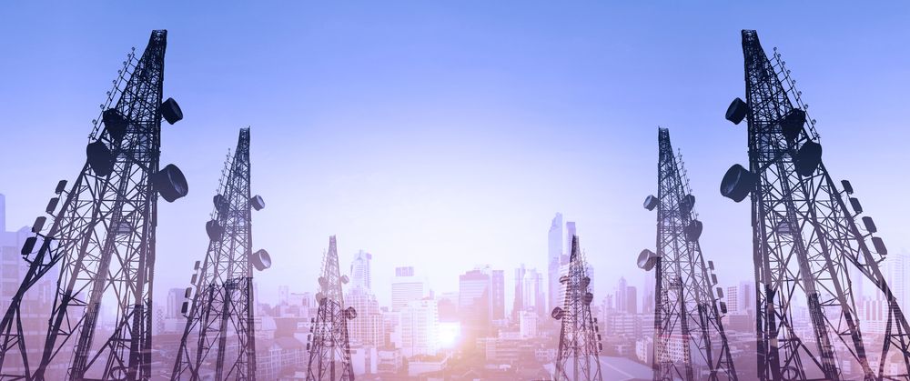Telecom masts loaded with antennas and dishes silhouetted over a city skyline at sunrise.